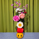 Colorful flower arrangement in a unique vase with a checkered tablecloth and green curtain background