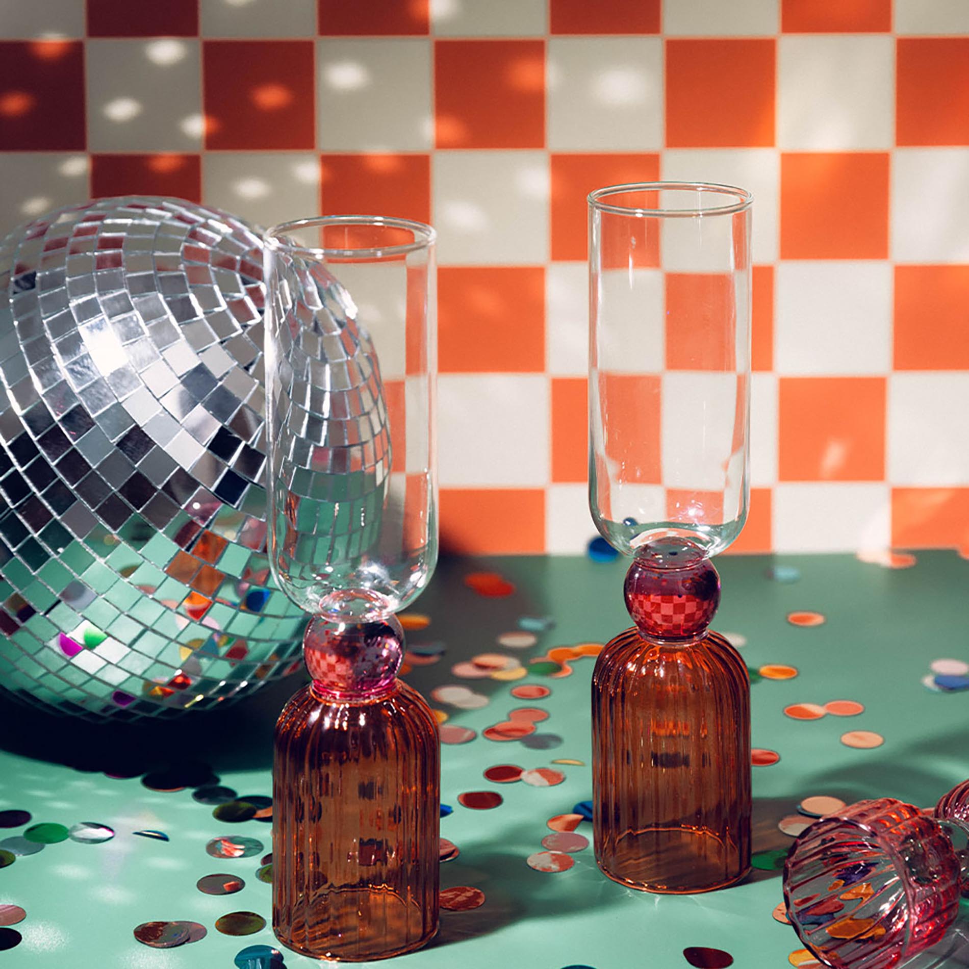 Two amber glass bottles with red stoppers and clear glasses on a checkered tablecloth with disco ball and confetti.