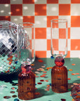 Two amber glass bottles with red stoppers and clear glasses on a checkered tablecloth with disco ball and confetti.