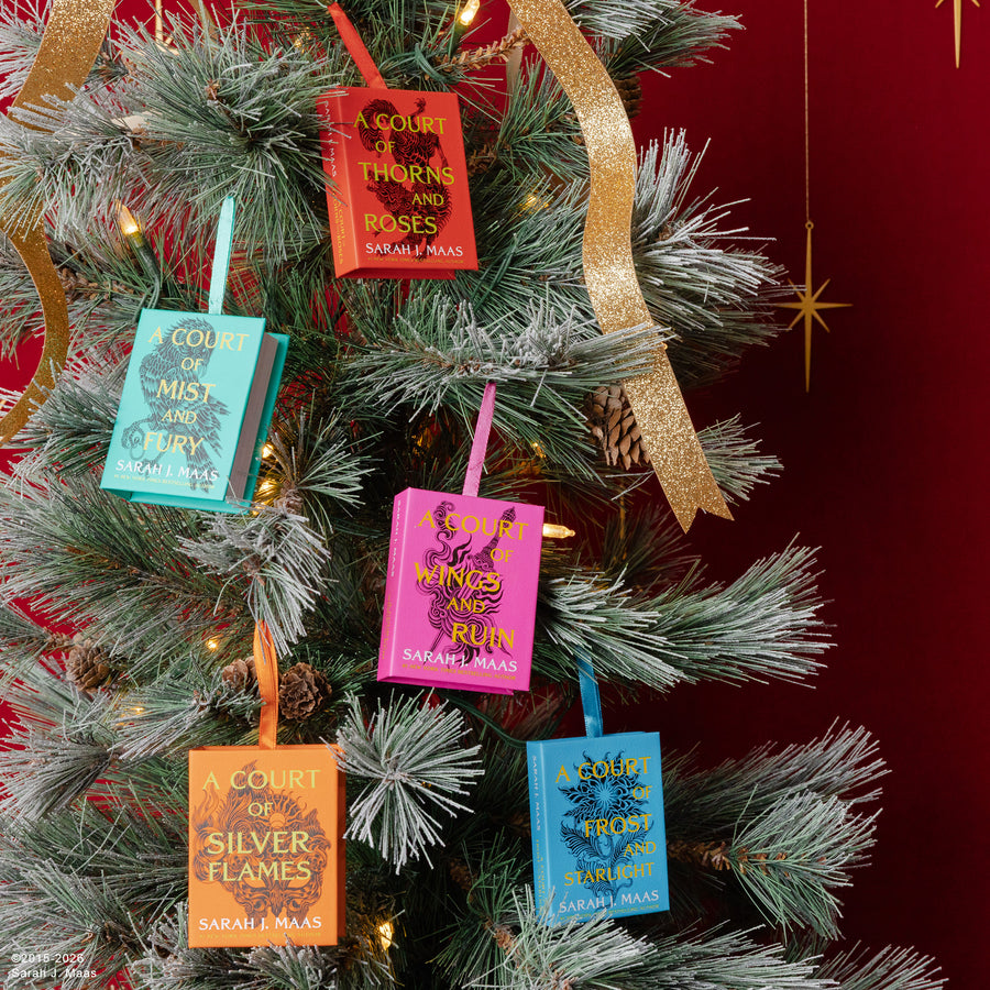 Six book-themed Christmas ornaments shaped like hardcover books hang from the branches of a decorated Christmas tree. The ornaments feature different book covers from Sarah Maas, including 'A Court of Mist and Mirrors', 'A Court of Thorns and Boots', 'A Court of Silver Flames', 'A Court of No Return', 'A Court of Blood and Ash', and 'A Court of Thorns and Boots' in vibrant colors like pink, orange, teal, and blue, with gold foil text.