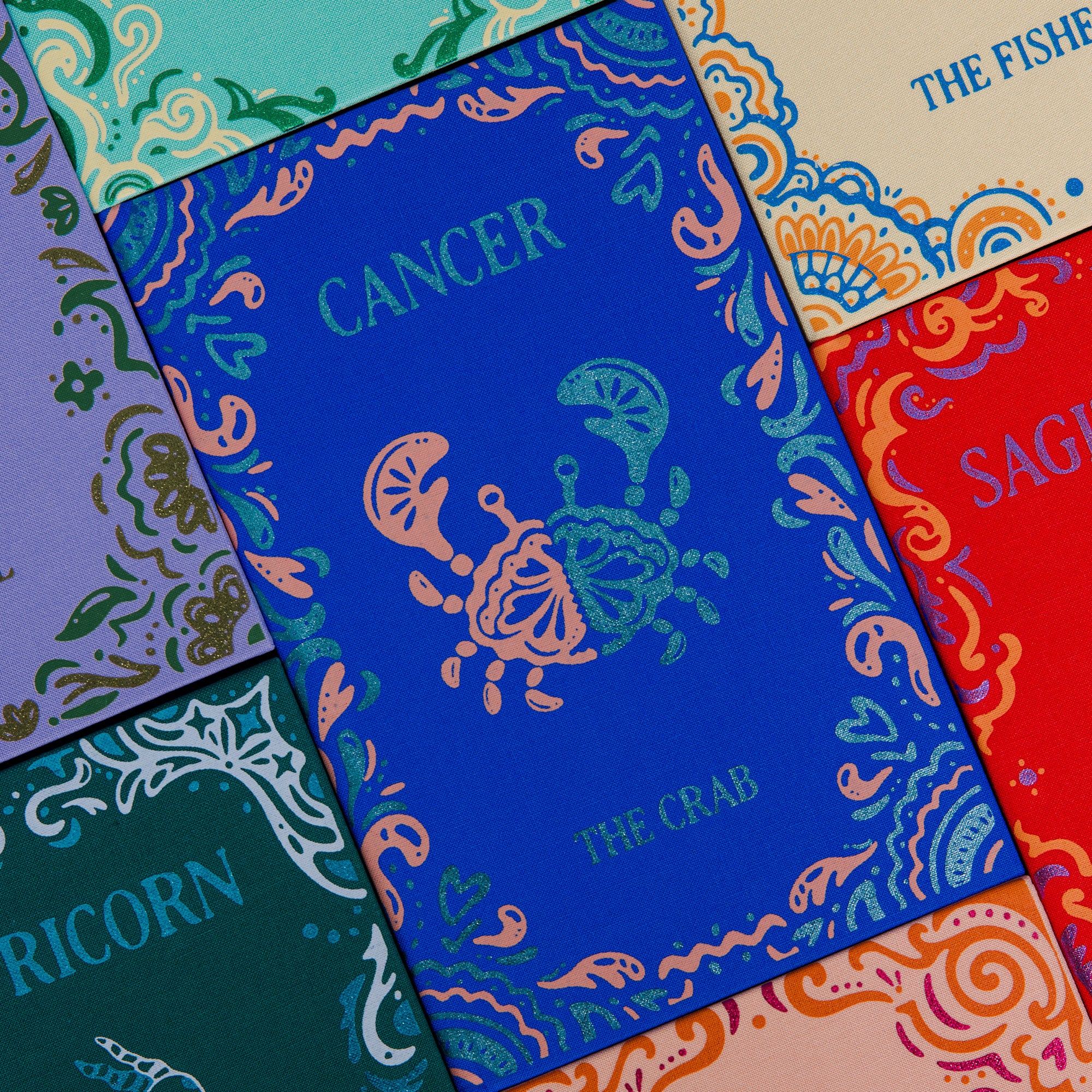 A set of colorful zodiac-themed cards or booklets arranged in a flat lay. The central card is blue with a teal crab illustration and the word 'CANCER'.