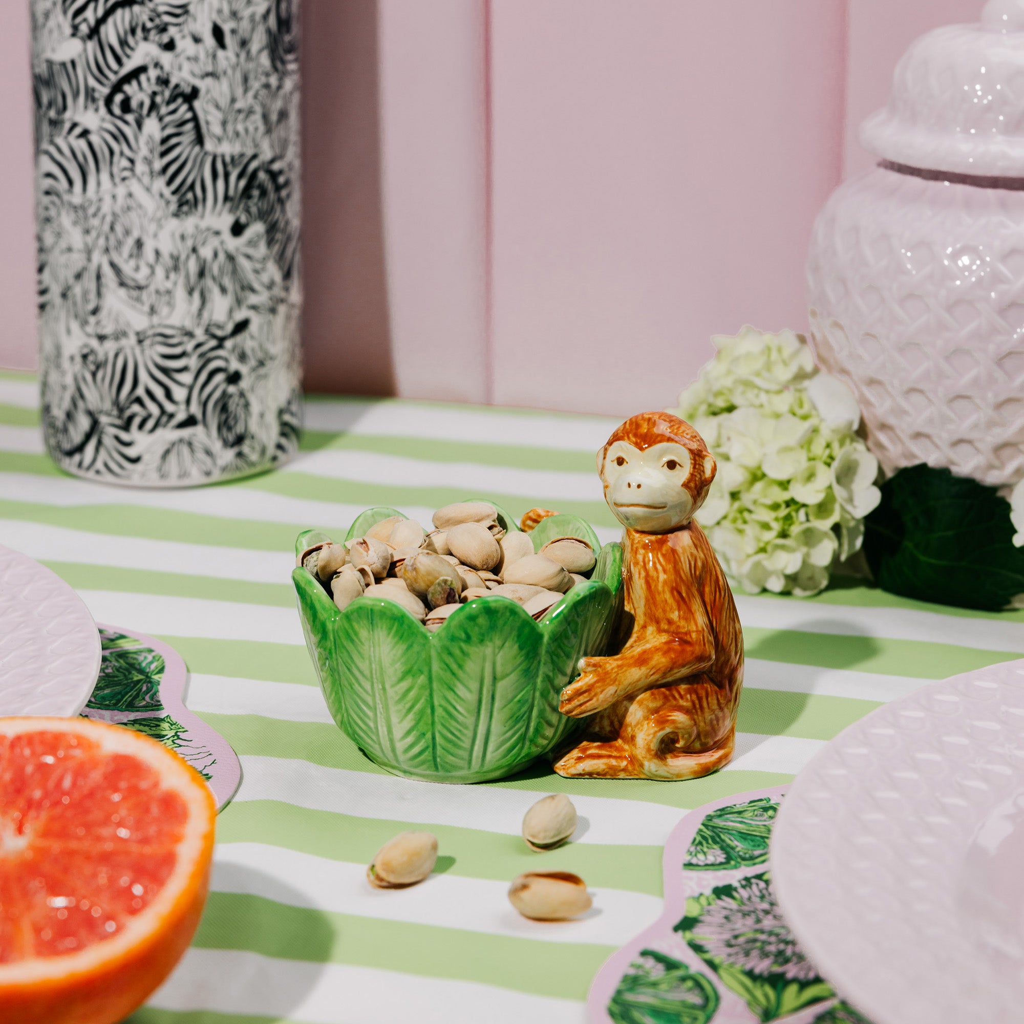 A green leaf-shaped ceramic bowl filled with pistachios sits beside a small orange-brown monkey figurine on a green and white striped tablecloth, styled with plates, a patterned vase, and fruit.