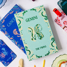 A mint green Gemini zodiac-themed notebook or card case with green and cream swirl patterns and twin illustrations, styled on a white surface with other colorful astrology-themed accessories.