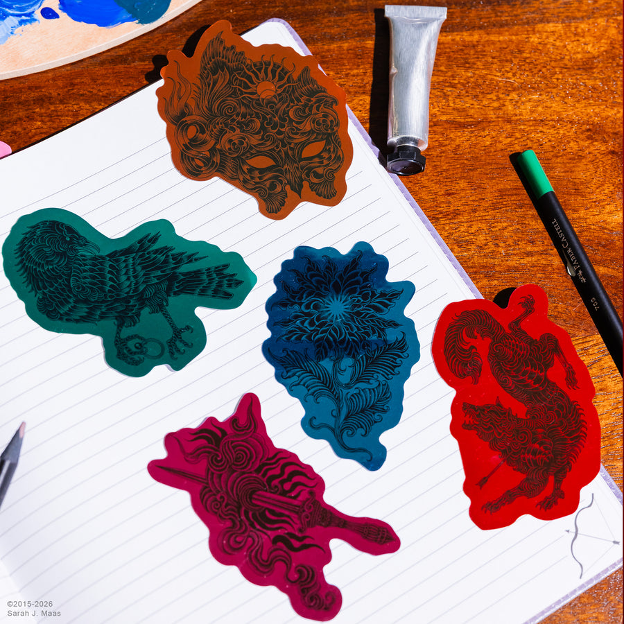 Five colorful stickers with intricate black line-art designs are arranged on a lined notebook page on a wooden desk, alongside an acrylic paint tube and a green pen.