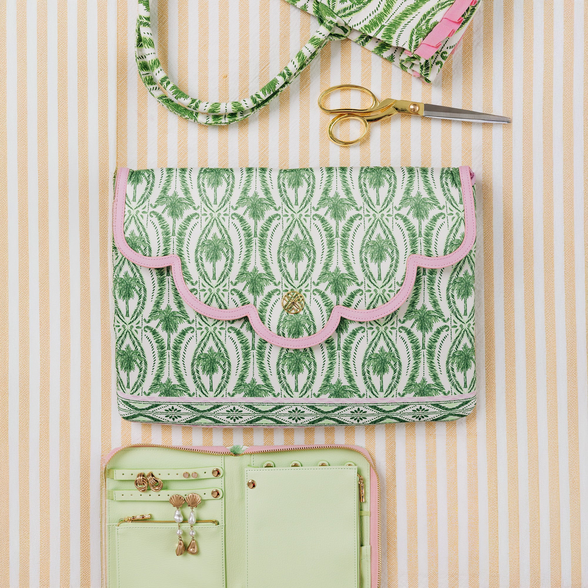 A green and white patterned fabric purse with a pink scalloped trim and a gold-tone clasp, displayed on a pink and white striped background with a matching fabric strap, gold scissors, and an open mint green jewelry organizer.