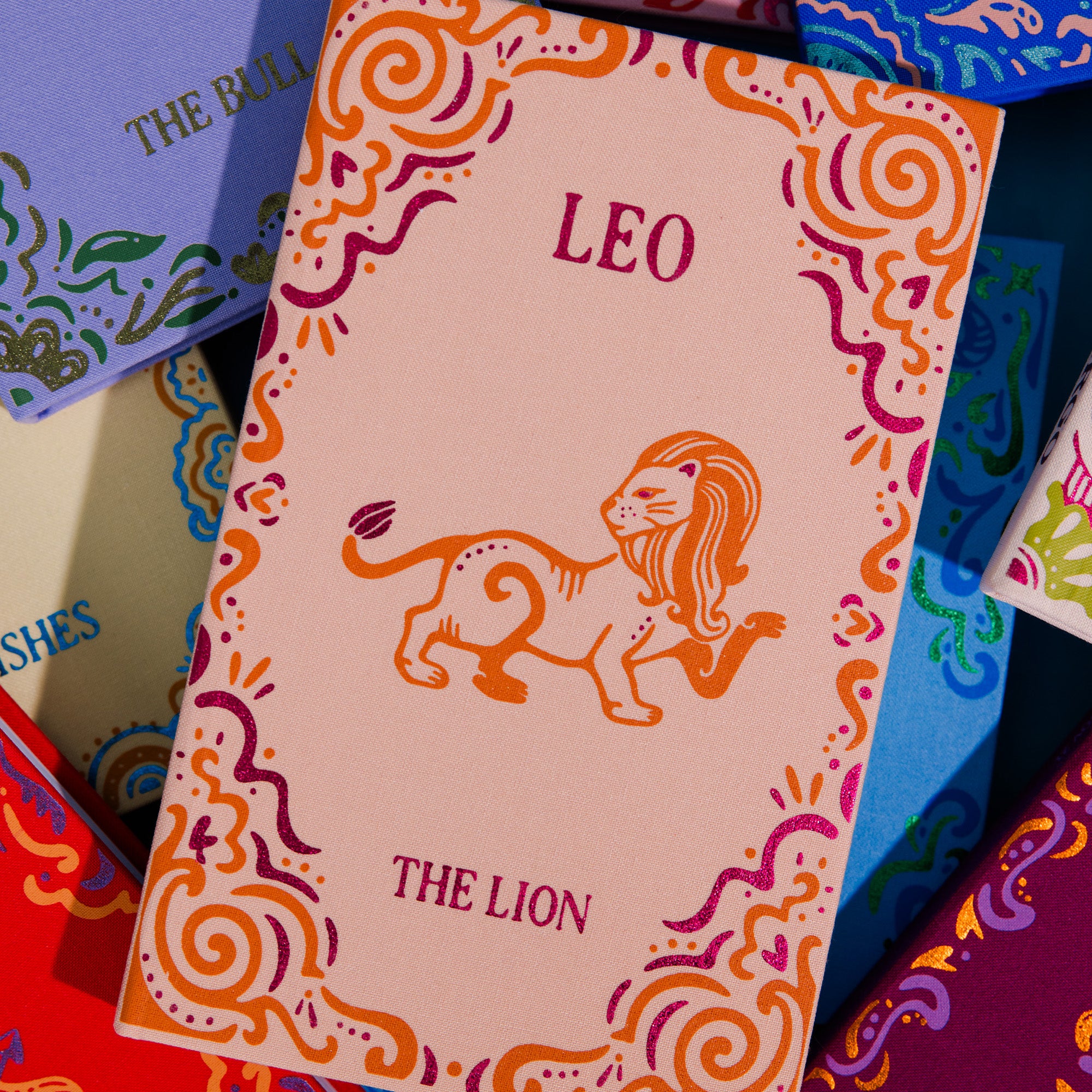 A close-up of a pink zodiac card titled 'LEO THE LION' with an orange lion illustration and decorative swirl borders, surrounded by other colorful patterned cards.