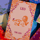 A close-up of a pink zodiac card titled 'LEO THE LION' with an orange lion illustration and decorative swirl borders, surrounded by other colorful patterned cards.