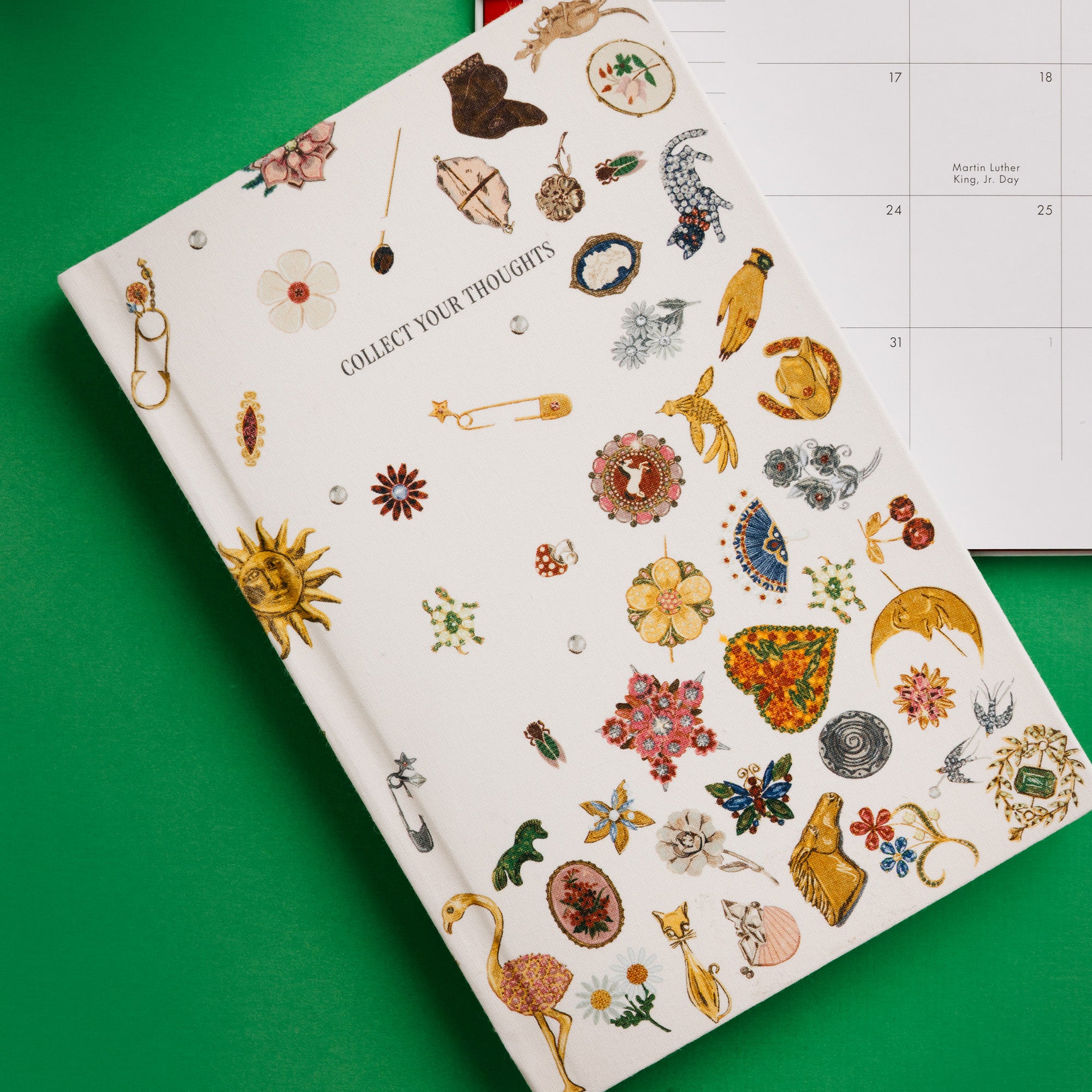 A cream-colored hardcover journal titled 'COLLECT YOUR THOUGHTS' decorated with various colorful vintage-style illustrations of jewelry, flowers, and symbols, shown on a bright green background with a monthly calendar partially visible behind it.