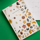 A cream-colored hardcover journal titled 'COLLECT YOUR THOUGHTS' decorated with various colorful vintage-style illustrations of jewelry, flowers, and symbols, shown on a bright green background with a monthly calendar partially visible behind it.