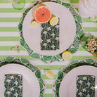 A styled flat-lay table setting featuring green and white striped plates with scalloped edges, patterned napkins, citrus slices, and pistachios on a green and white striped tablecloth.