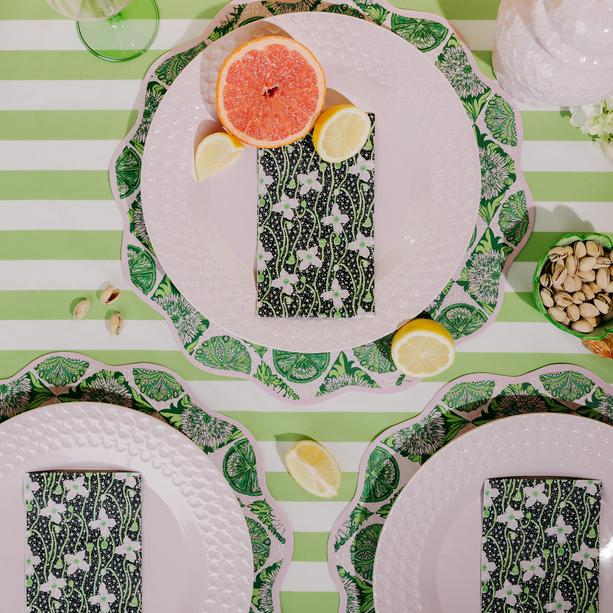 A styled flat-lay table setting featuring green and white striped plates with scalloped edges, patterned napkins, citrus slices, and pistachios on a green and white striped tablecloth.