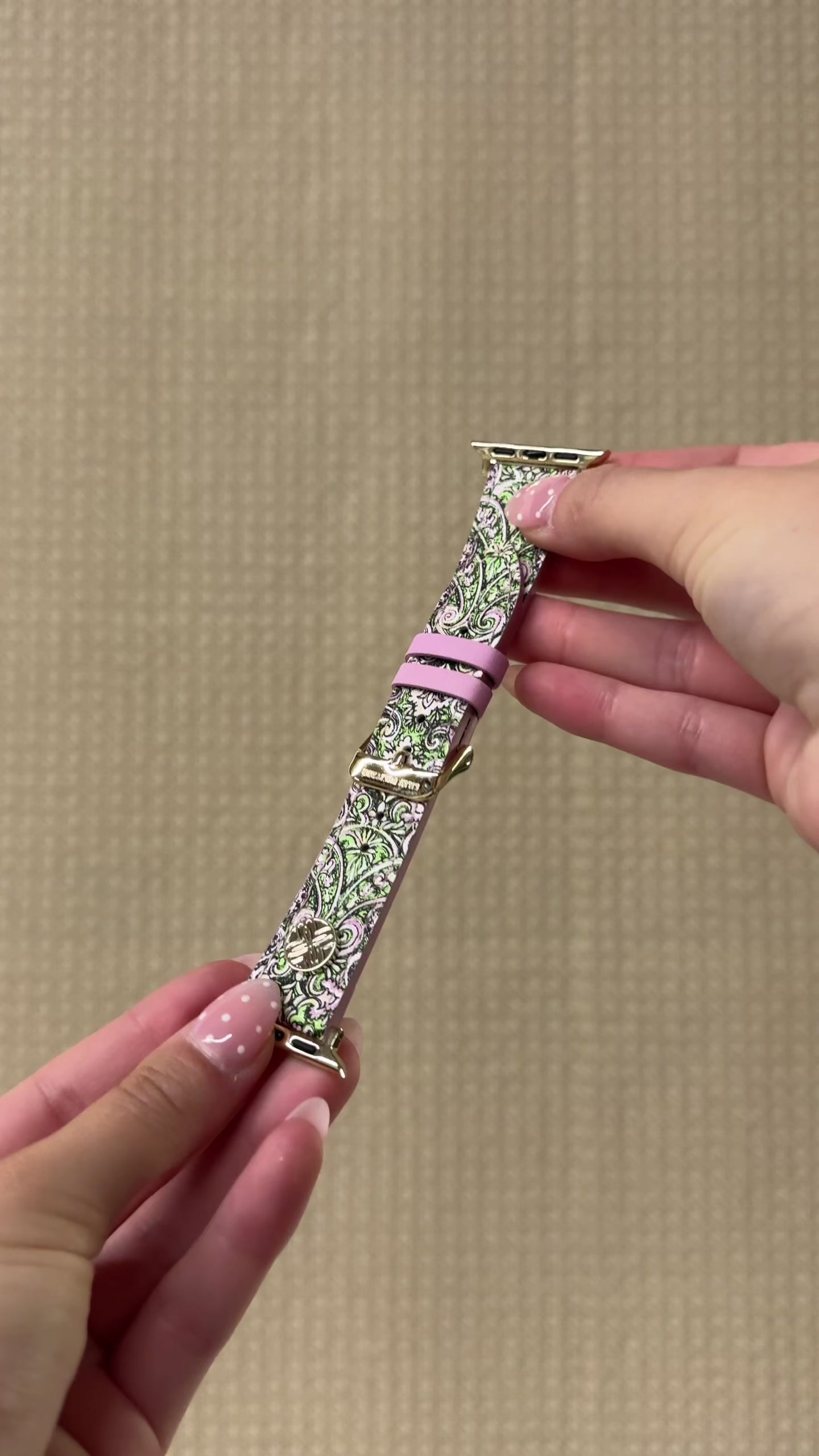 model showcasing a smartwatch with a black rectangular screen and a patterned strap featuring green, pink, and white floral motifs with a gold buckle, shown against a plain white background.