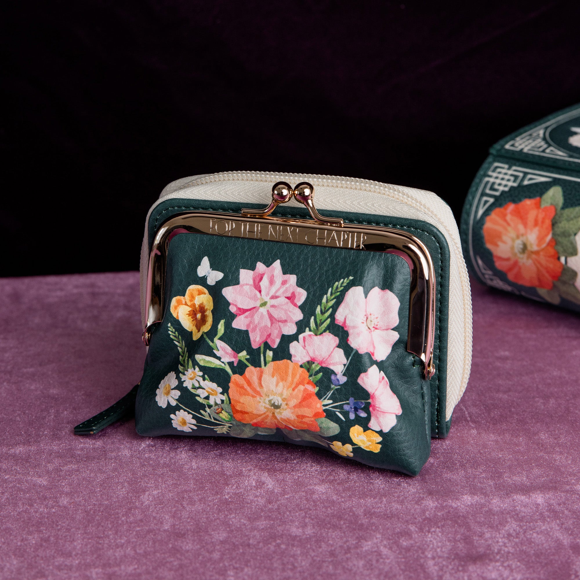A small floral coin purse with a dark green base, colorful flower print, cream zipper, and gold-tone kiss-lock closure, displayed on a purple velvet surface with a dark background.