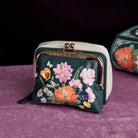 A small floral coin purse with a dark green base, colorful flower print, cream zipper, and gold-tone kiss-lock closure, displayed on a purple velvet surface with a dark background.