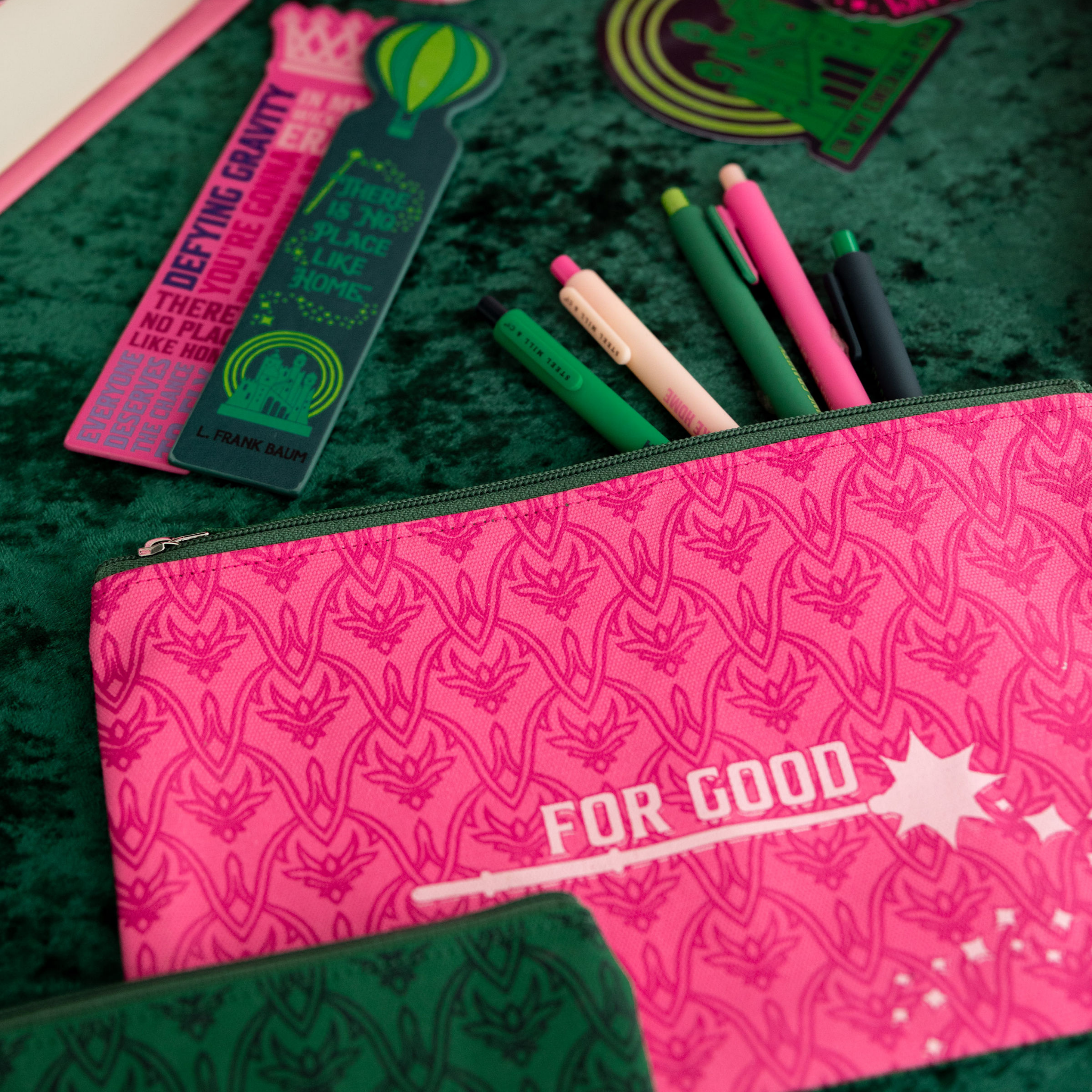 Pink pencil case with 'FOR GOOD' text on a green textured surface with stationery items.