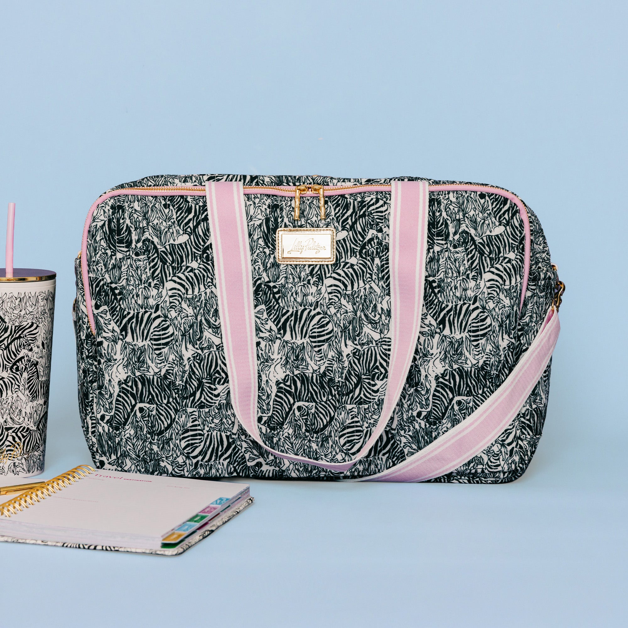 A rectangular duffel bag with a black and white zebra print, pink velvet handles, and a gold-tone brand plaque. It is styled on a light blue background next to a matching zebra-print tumbler and notebooks.