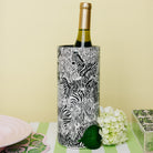 A wine bottle with a gold foil top stands inside a cylindrical holder covered in a black-and-white zebra print. The holder sits on a green-and-white striped surface with a pale yellow background, white flowers, and partial decorative objects at the edges.